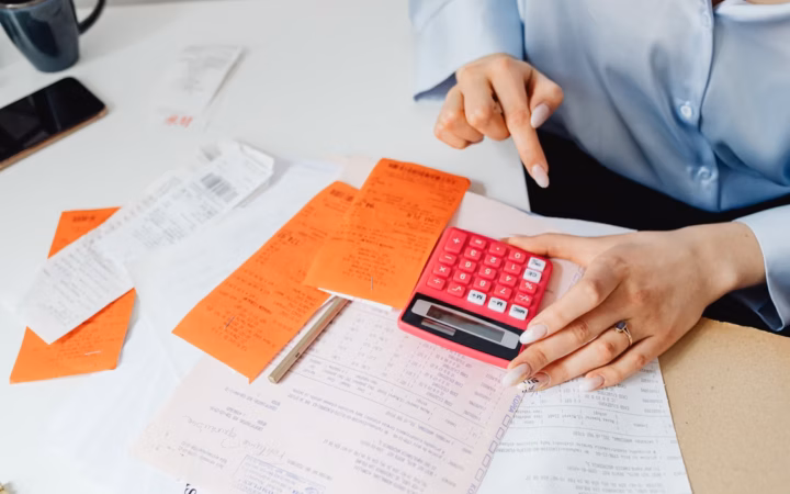 Mano utilizando una calculadora junto a documentos y una tabla de gastos; ilustración editorial, no representa un informe oficial de fiscalización.