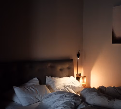 Dormitorio en penumbra con una lámpara encendida y una cama preparada para la noche.