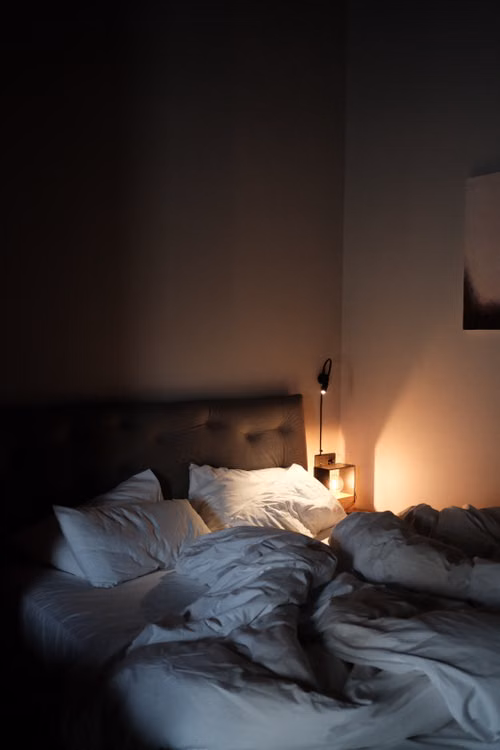 Dormitorio en penumbra con una lámpara encendida y una cama preparada para la noche.